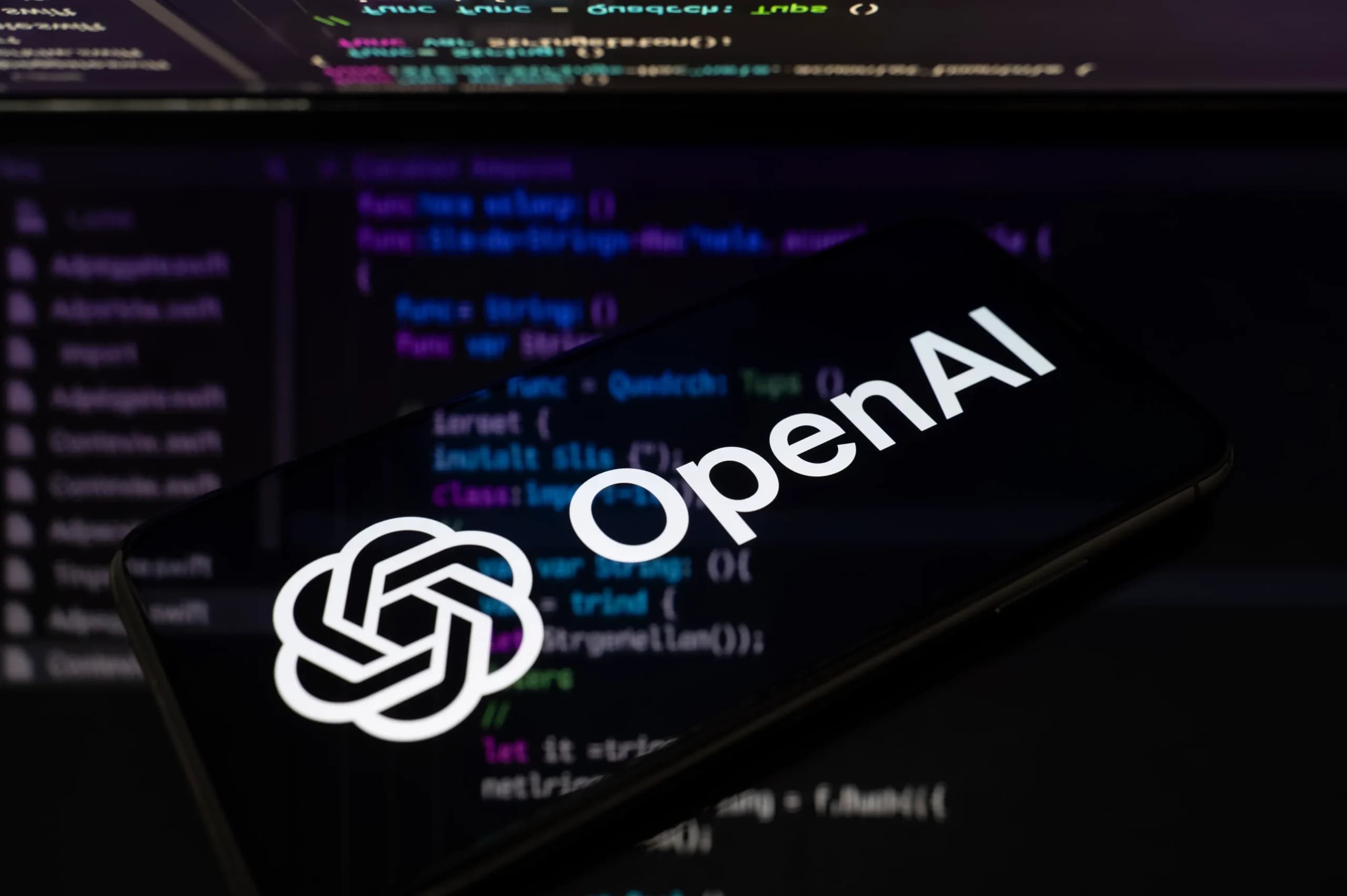 Logotipo da OpenAI sobre a tela de um smartphone com código ao fundo